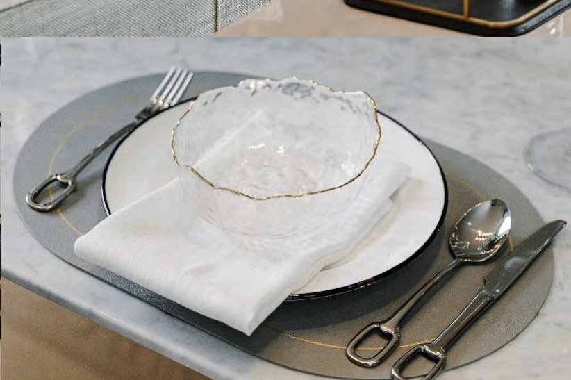 ภาพระยะใกล้ของการจัดโต๊ะรายที่นั่ง ชามแก้วขอบทองผิวลายนูน ผ้าเช็ดปากลินินพับ จานรอง และช้อนส้อมขัดเงาบนหินอ่อนสีขาว