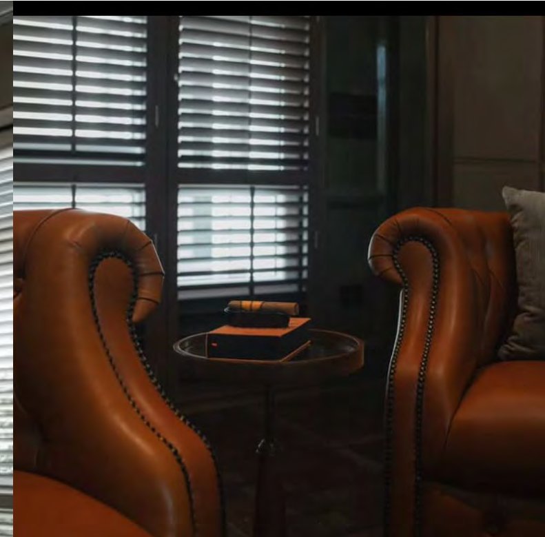 เก้าอี้ปีก Chesterfield หนังสีแทนตอกหมุดสองตัว หันเข้าหากันคนละฝั่งของโต๊ะทรงกลมตัวเล็กที่วางกล่องซิการ์ไว้ พร้อมบานเกล็ด plantation ด้านหลัง