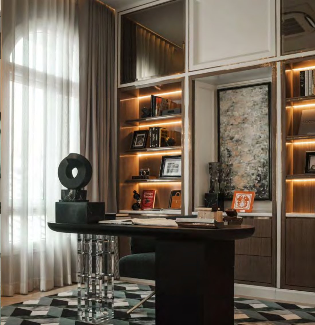 มุมห้องทำงานในบ้าน โต๊ะทรงรีสีเข้มบนขาคริสตัล ชั้นหนังสือที่มีไฟส่องจากด้านหลัง ภาพแอบสแตรกต์ใส่กรอบ และพรมสีเขียวลายเรขาคณิต