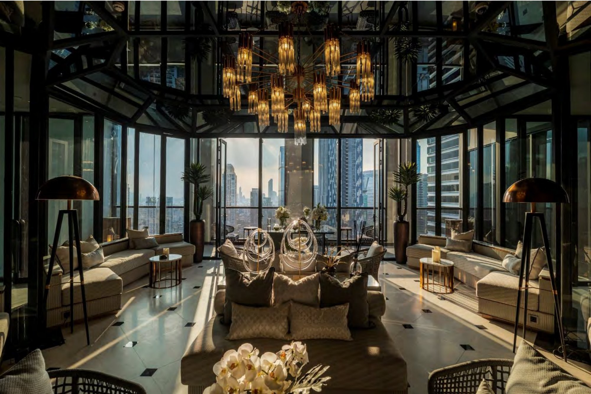 sky lounge เพดานสูงพร้อมโคมระย้าคริสตัลไหลลดหลั่น โซฟาโทนอ่อน โคมไฟขาตั้งสามขา และเส้นขอบฟ้ากรุงเทพที่มองผ่านผนังกระจกทรงเหลี่ยม
