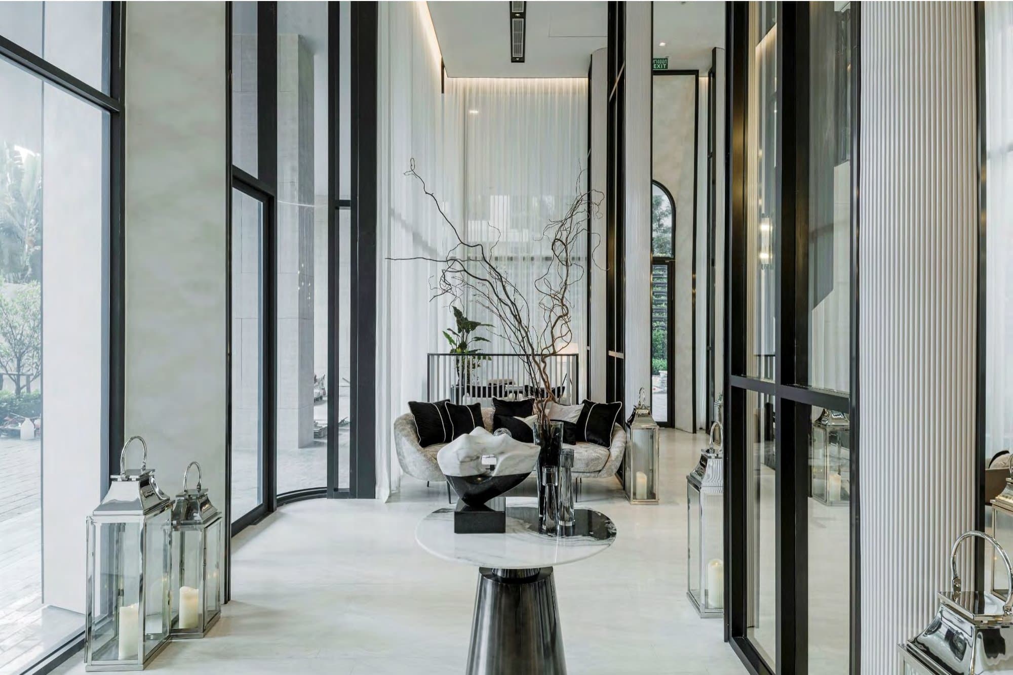 Double-height arrival lobby with black steel-framed glazing, pale marble floor, polished silver lanterns and a bouclé sofa beneath sculptural bare branches